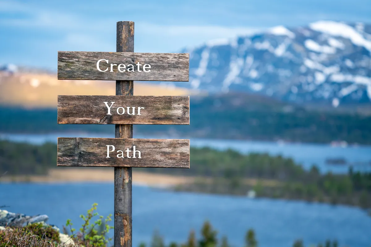 Holzwegweiser mit der Aufschrift „Create Your Path“ vor einer Berglandschaft
